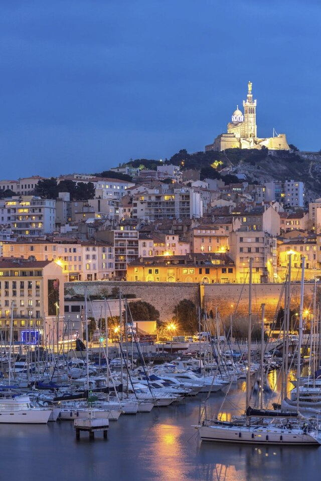 Vue nocturne du Vieux-Port de Marseille avec Notre-Dame de la Garde illuminée en arrière-plan.